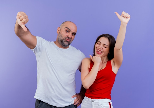 Adult couple confident man keeping hand on waist and joyful woman with closed eyes both showing thumbs up 