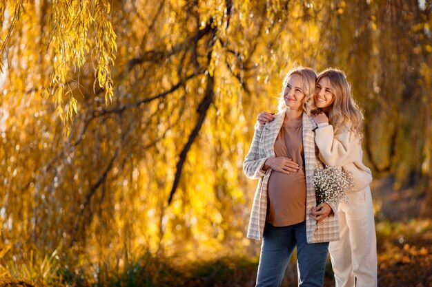 성인 딸은 자연 속에서 화창한 가을날 임신한 엄마를 안고 있습니다. 그녀의 손에 꽃을 들고 있는 금발의 여자 베이지색 옷을 입은 여성
