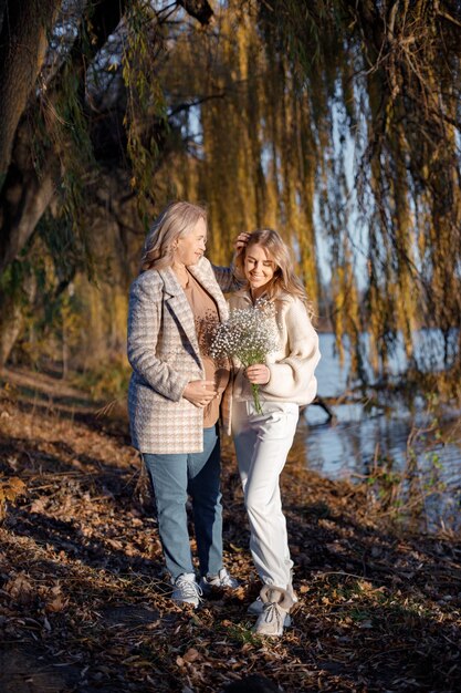 Adult daughter hugs her pregnant mom on a sunny autumn day in nature Blonde woman holding flowers in her hand Women wearing beige clothes