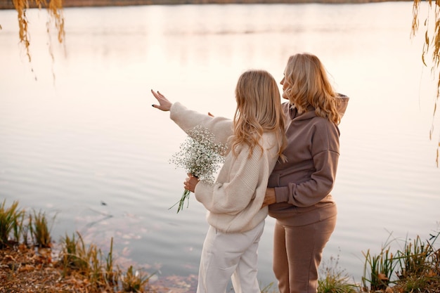 성인 딸은 자연 속에서 화창한 가을날 임신한 엄마를 안고 있습니다. 호수 근처 공원에 서 있는 금발 여성 베이지색 옷을 입은 여성