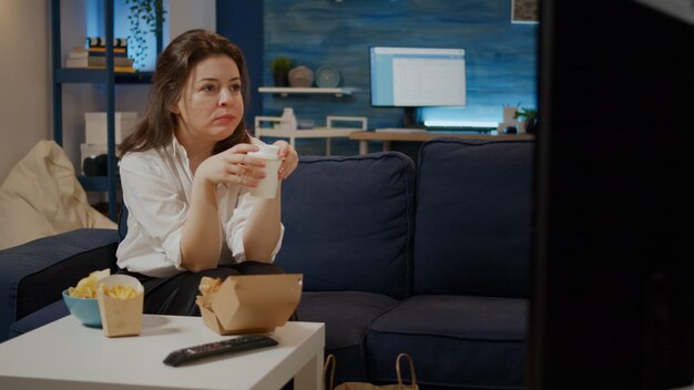 Adult eating hamburger and fries watching television in living room sitting on couch. Young woman with fast food meal enjoy eating and drinking soda at home from takeaway delivery place