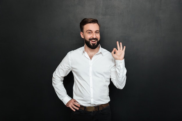 Adult guy in office posing on camera, smiling and gesturing with OK sign expressing everything is alright over dark gray