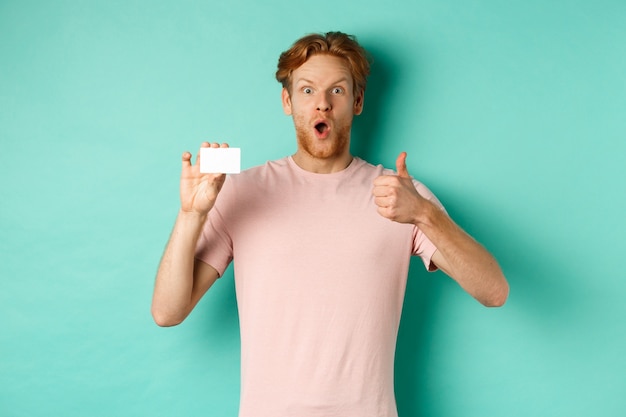 Adult guy with red hair and beard showing plastic credit card and thumb up, looking impressed, recommend bank, standing over mint background