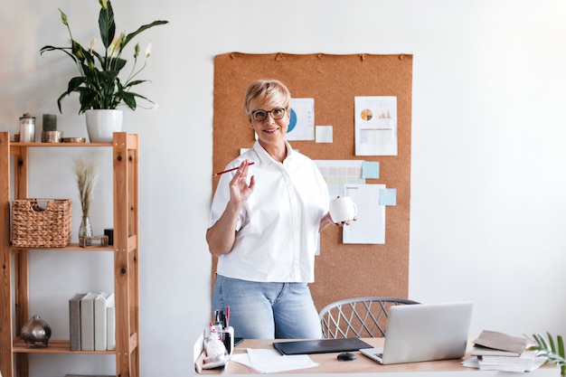 Adult lady in good mood holds coffee cup and poses in office