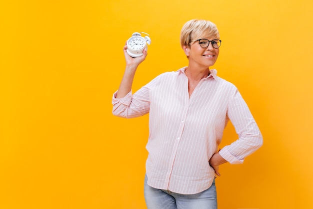 Adult lady looks into camera and demonstrates alarm clock on orange background