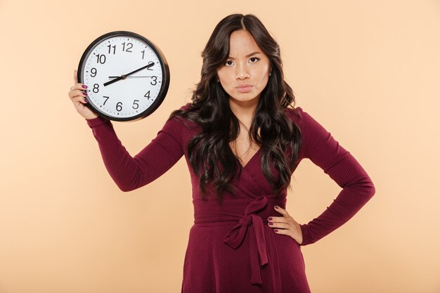 Adult lady with curly long hair holding clock with time after 8 showing anger with facial expressions putting hand on waist over beige background