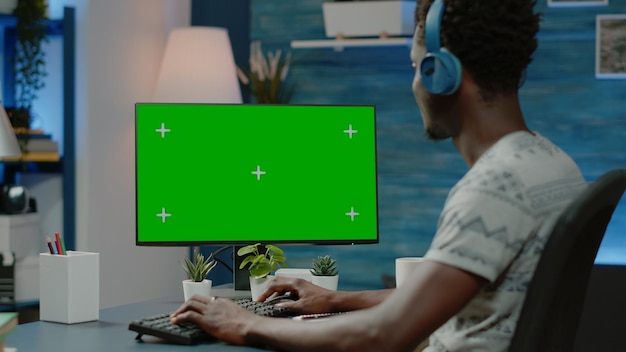 Adult looking at horizontal green screen on computer for work. Employee with headphones working from home with chroma key on device for mockup template and isolated background.