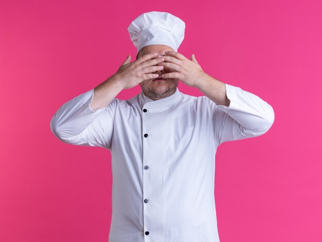 взрослый мужчина-повар в униформе шеф-повара и очках держит руки перед глазами, изолированными на розовой стене