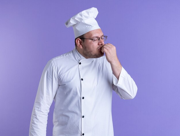 взрослый мужчина-повар в униформе шеф-повара и очках, касаясь губ рукой с закрытыми глазами, изолированными на фиолетовой стене