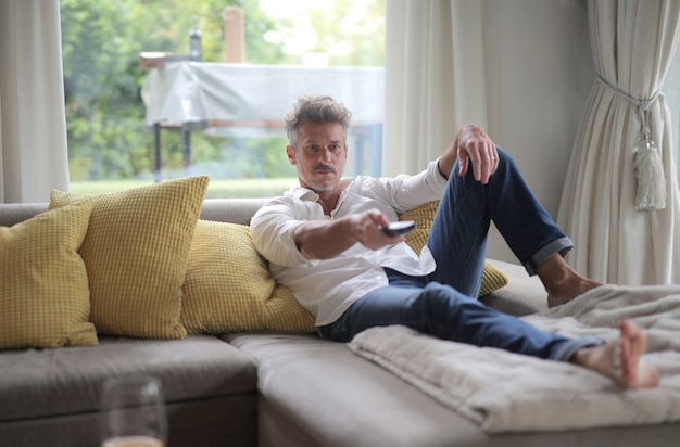 Adult male lying on the sofa and holding a remote control under the sunlight through the windows