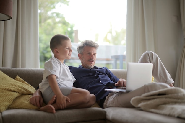 Adult male lying on the sofa with his son and using the laptop under the lights