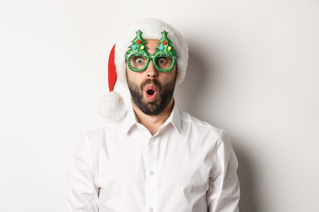 Foto gratuita uomo adulto che celebra le vacanze invernali, indossando occhiali da festa di natale e cappello da babbo natale, guardando sorpreso dalla fotocamera, in piedi