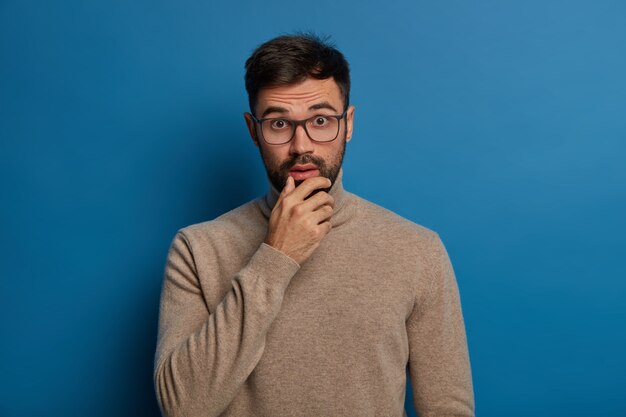 Adult man holds chin, looks with shock at camera, has embarrassed expression, reacts on unexpected news, wears transparent glasses and casual jumper, stands against blue wall.