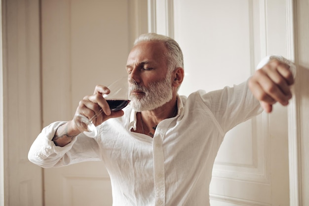 흰 옷을 입은 성인 남자는 와인을 즐긴다 밝은 셔츠에 회색 머리를 한 수염 난 남자가 빨간 음료를 마시고 카메라에 포즈를 취하고 있다