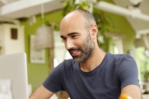 Adult man with beard dressed casually using modern laptop computer, checking email or messaging online, sitting against green interior.