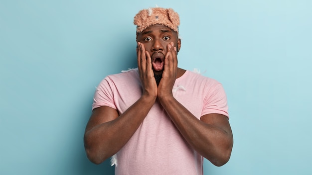 Free photo adult man with dark skin, feels shocked after having not enough sleep, red eyes, wakes up too early, wears eyemask, casual t shirt, isolated over blue wall. stunned afro man in nightwear
