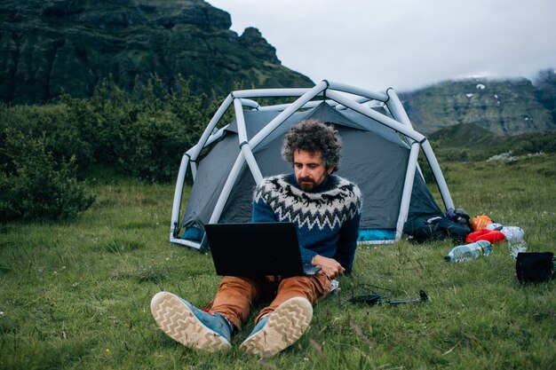Взрослый мужчина с седой бородой и забавными вьющимися хипстерскими волосами сидит перед палаткой на траве, удаленно работает с ноутбуком