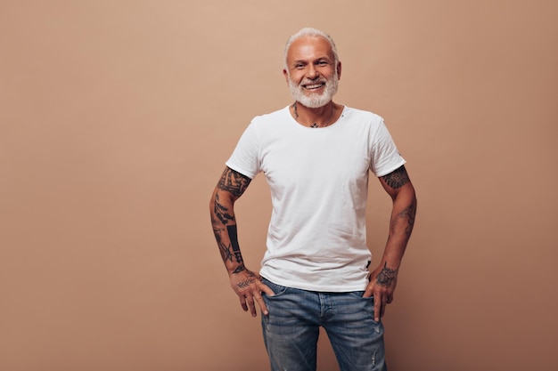 Adult man with tattoos poses on beige background Handsome guy with gray beard in white tshirt and blue modern jeans smiles
