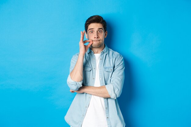 Adult man zipping mouth, promise keep secret, making a seal on lips and standing over blue background