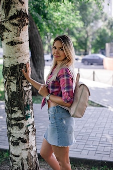 Adult shining blonde woman with face looking into camera standing in park near birch wearing checker...