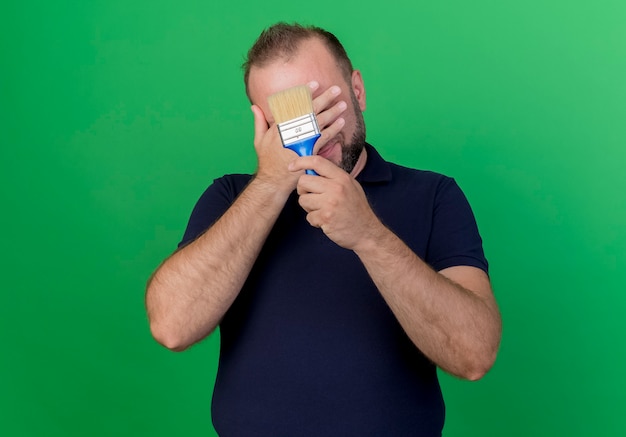 Adult slavic man holding paint brush covering face with hand isolated on green wall