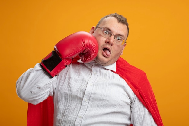 Adult slavic superhero man in red cape wearing glasses and box gloves looking at side beating himself in face with tongue pulled out isolated on orange wall