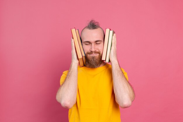 Взрослый улыбающийся парень в повседневной одежде с бородой обнимает себе любимые книги, изолированные на розовом