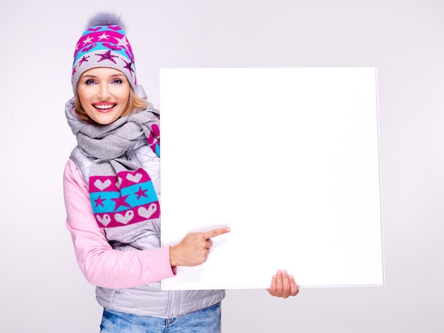 Adult smiling woman in warm outerwear holds the white placard in hands and points on it