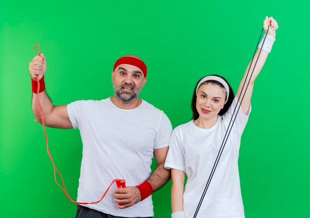 Adult sporty couple wearing headband and wristbands impressed man holding jump rope and pleased woman pulling jump rope both looking 