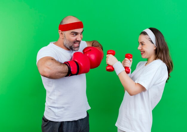 Adult sporty couple wearing headband and wristbands impressed man wearing box gloves keeping hands together smiling woman holding dumbbells looking 