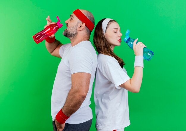 Adult sporty couple wearing headband and wristbands standing back to back drinking water from bottle 