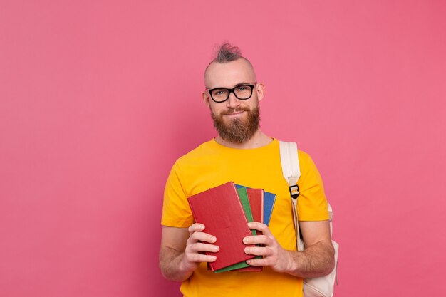 Взрослый студент веселый парень повседневной одежды с бородой и рюкзаком держит книги, изолированные на розовом