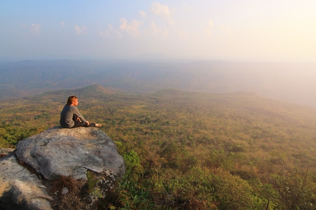 Adult tourist in black trousers, jacket and dark cap sit on cliff's edge and looking to misty hilly valley bellow