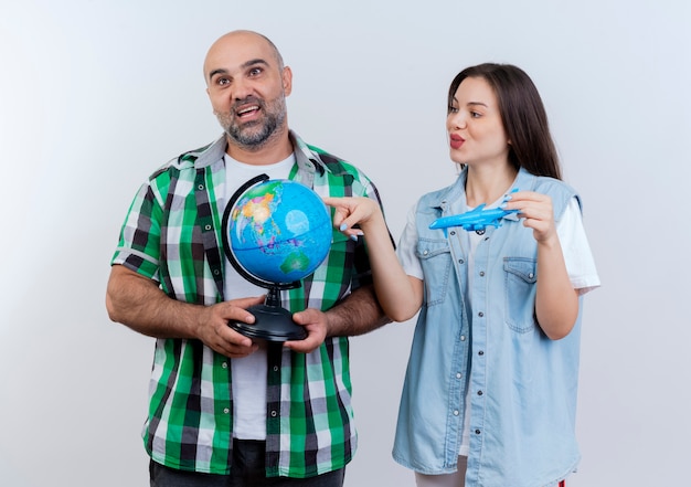 Adult traveler couple impressed man holding globe looking straight and pleased woman holding model plane looking at globe and touching it 