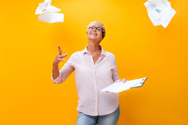 Adult woman in eyeglasses scatters paper sheets on orange background