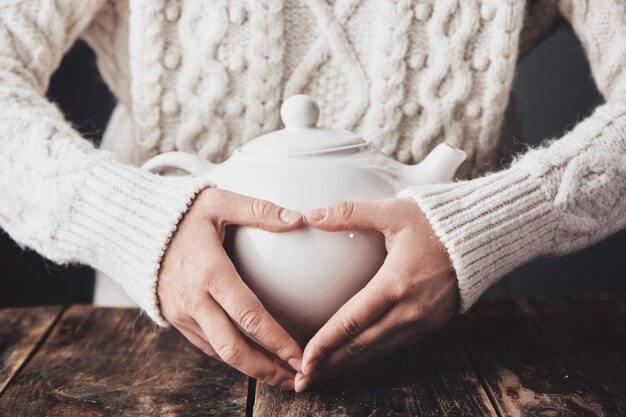 Руки взрослой женщины обнимают большой керамический чайник с горячим напитком внутри.