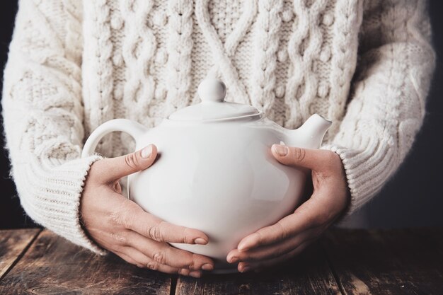 Adult woman hands hug big ceramic teapot with hot drink inside.