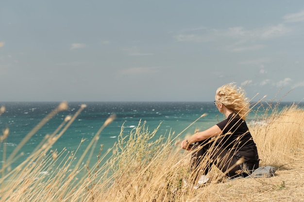 An adult woman sits on a high bank overlooking the ocean her hair blowing in the wind