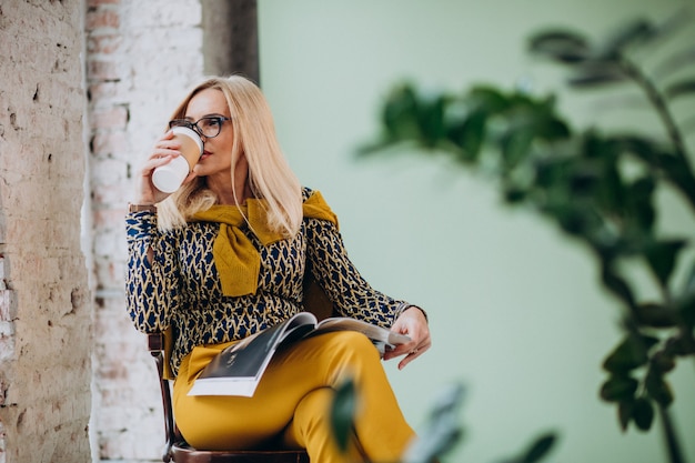 Donna adulta che si siede nella sedia che beve caffè e che legge rivista