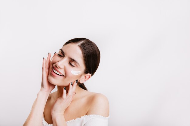 Adult woman in white top cares for skin of face, applying cream. Portrait of girl with ponytail on isolated wall.