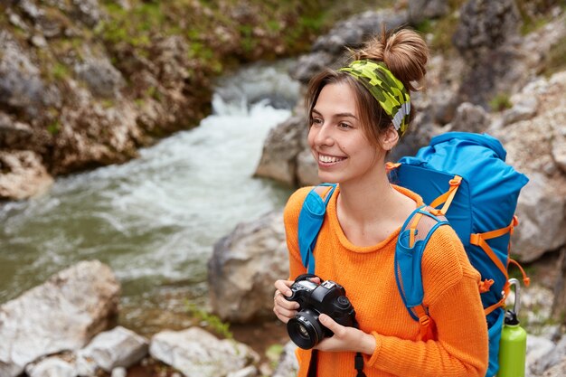 Adventurous female enjoys travel adventure, has hiking activity and nature tour, makes photo of landscapes, holds professional camera