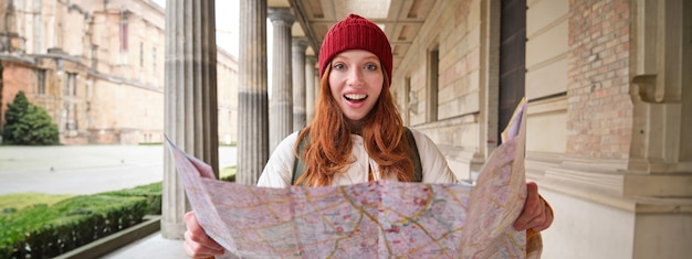 Free photo adventurous redhead girl walks in town with paper map explores city as tourist looks for popular