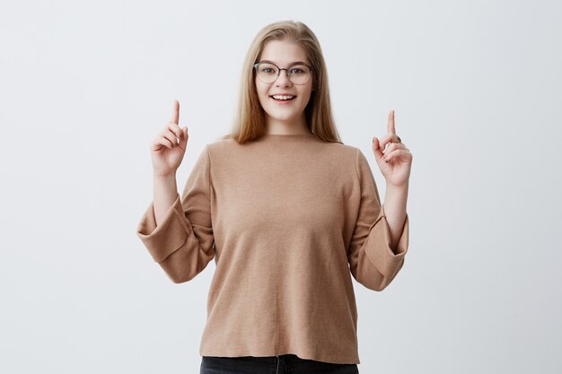 Advertisment concept. Happy woman in sweater, points up with fore fingers as shows something, has surprised pleased expression. Blonde female wearing stylish eyewear gestures against gray wall