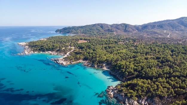 Побережье Эгейского моря с голубой прозрачной водой, зеленью вокруг, скалами, кустами и деревьями, вид с дрона, Греция