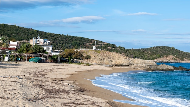 Aegean sea coast with buildings on the left, rocks, bushes and trees, blue water with waves in Sarti, Greece