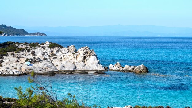 Побережье Эгейского моря с зеленью вокруг, скалами и кустами, голубой водой и отдыхающими людьми, Греция