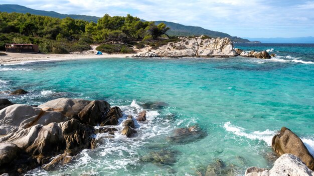 주변의 녹지, 바위, 관목 및 나무, 파도가있는 푸른 물, 그리스가있는에게 해 해안