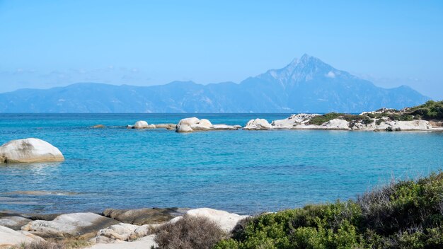 Побережье Эгейского моря со скалами над водой и сушей вдалеке, зелень на переднем плане, голубая вода, Греция