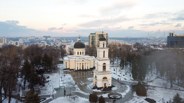 Aerial drone view of Chisinau downtown in winter. Panorama view of central park with snow, trees and multiple walking people, belltower, cathedral, buildings on the background.