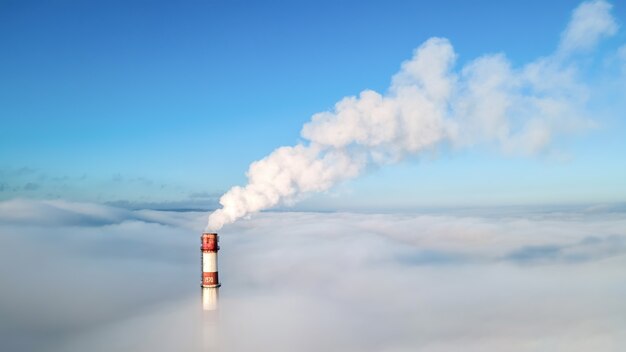 Aerial drone view of thermal station's tube visible above the clouds with smoke coming out. Blue and clear sky